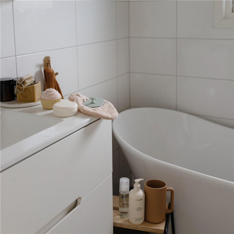 Mushie Silicone Baby Bath Cradle Cap Brush in Tradewinds & Periwinkle with bath accessories on a vanity near a bathtub.