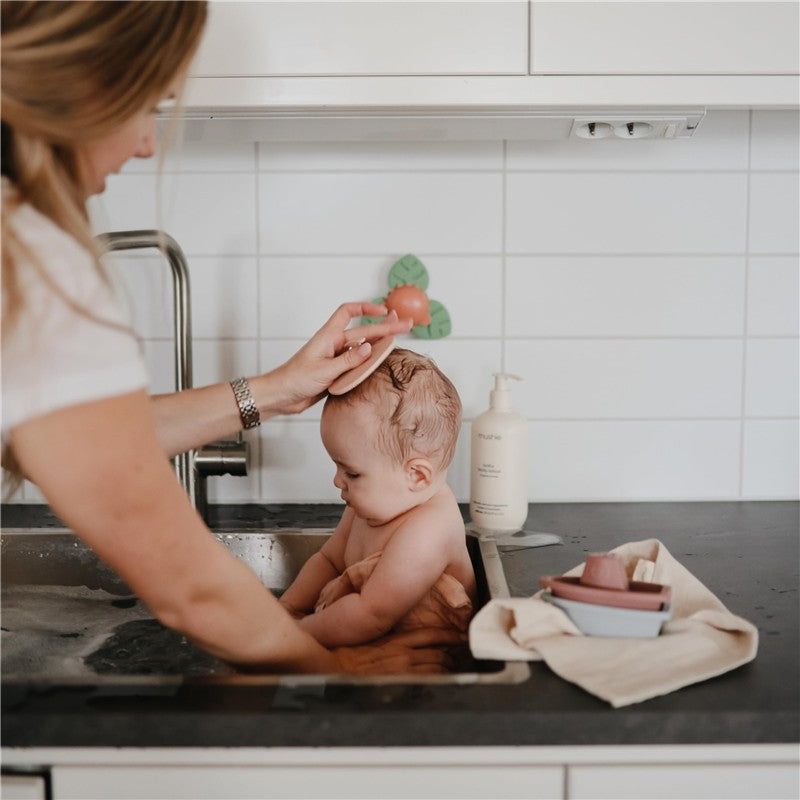 A parent uses a Mushie silicone cradle cap brush on a baby during bath time, with toys visible in the sink.
