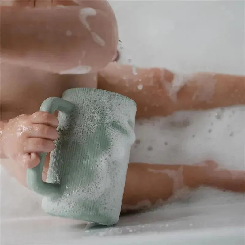 Sage green Mushie bath rinse cup held by a child in a bubbly bath, illustrating its use for rinsing.