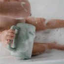 Sage green Mushie bath rinse cup held by a child in a bubbly bath, illustrating its use for rinsing.