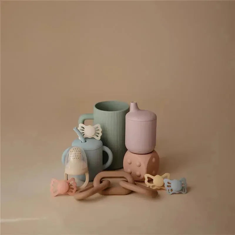 Mushie Bath Rinse Cup in sage, accompanied by pastel-colored cups and silicone toys arranged aesthetically.