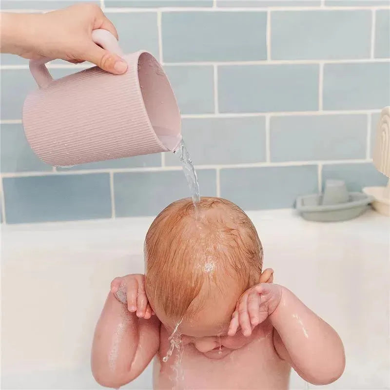A hand pours water from a sage-colored Mushie bath rinse cup over a baby’s head in a bathtub.