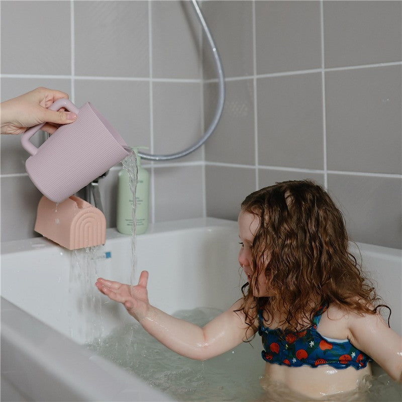 Mushie Bath Rinse Cup in blush color being used to pour water over a smiling child in a bathtub.