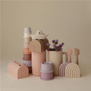 Mushie Bath Rinse Cup in blush, surrounded by various pastel-colored silicone containers and utensils.