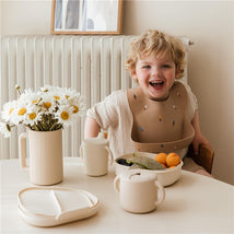 Smiling child wearing a patterned bib, sitting at a table with a Mushie bath rinse cup and a bowl of fruit.