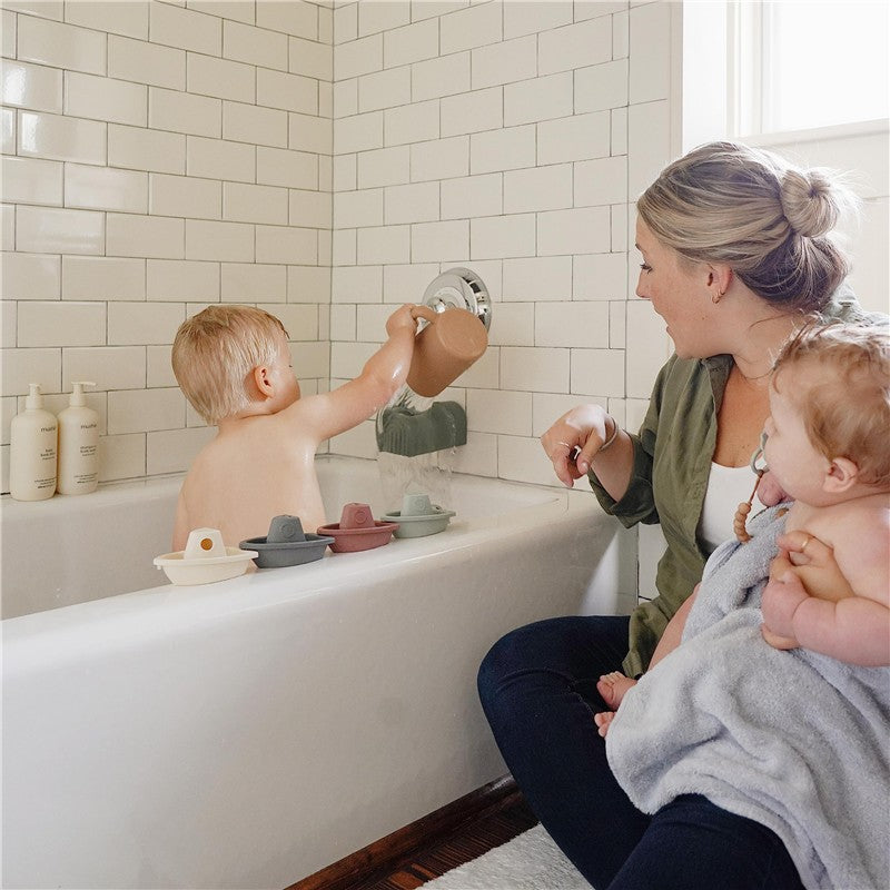 A child using the Mushie Bath Rinse Cup in a bathtub, with a parent sitting nearby holding another child.