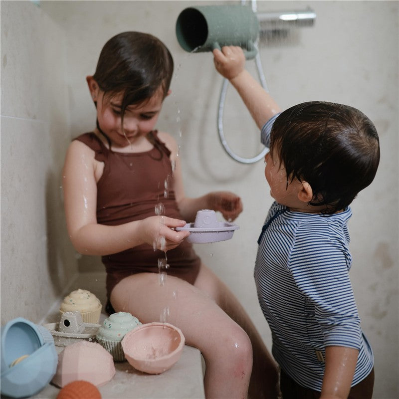 A child with short hair plays with a pink bath rinse cup in a shower, while another child pours water from a green rinse cup.