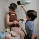A child with short hair plays with a pink bath rinse cup in a shower, while another child pours water from a green rinse cup.