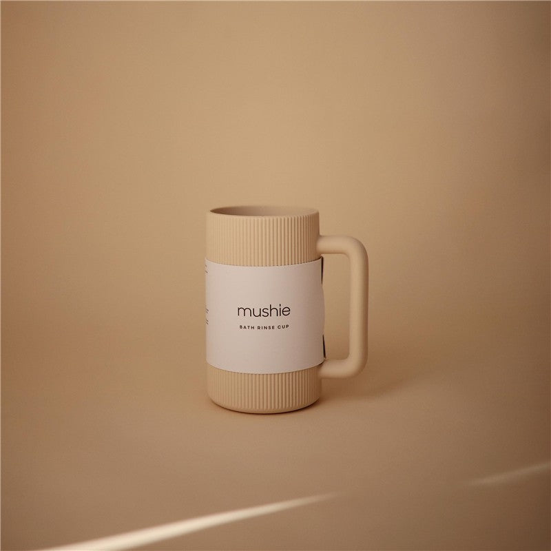 Mushie bath rinse cup in shifting sand color, featuring ribbed design and a white label with brand name and product details.