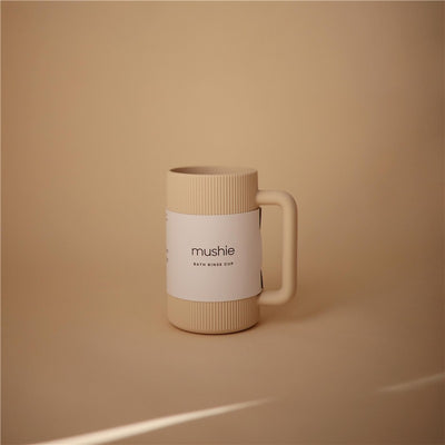 Mushie bath rinse cup in shifting sand color, featuring ribbed design and a white label with brand name and product details.