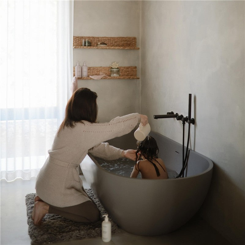 A woman gently rinses a child’s hair with a white cup while kneeling beside a modern gray bathtub in a serene bathroom.