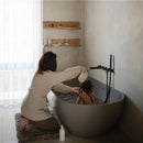 A woman gently rinses a child’s hair with a white cup while kneeling beside a modern gray bathtub in a serene bathroom.