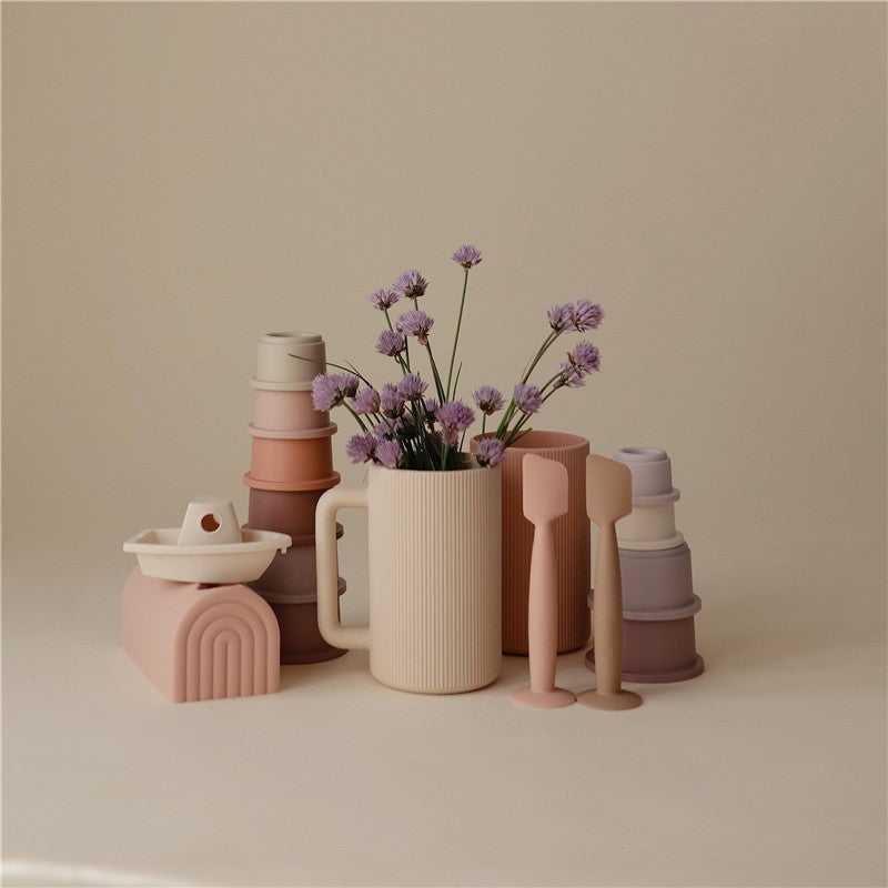Mushie Bath Rinse Cup in Shifting Sand color, surrounded by stacking cups and a flower arrangement.