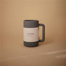 Gray Mushie bath rinse cup with a textured handle and branding label, placed on a beige background.