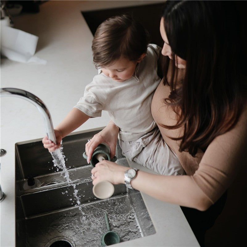 A woman and a child are washing a Mushie silicone baby bottle brush in a sink with running water.