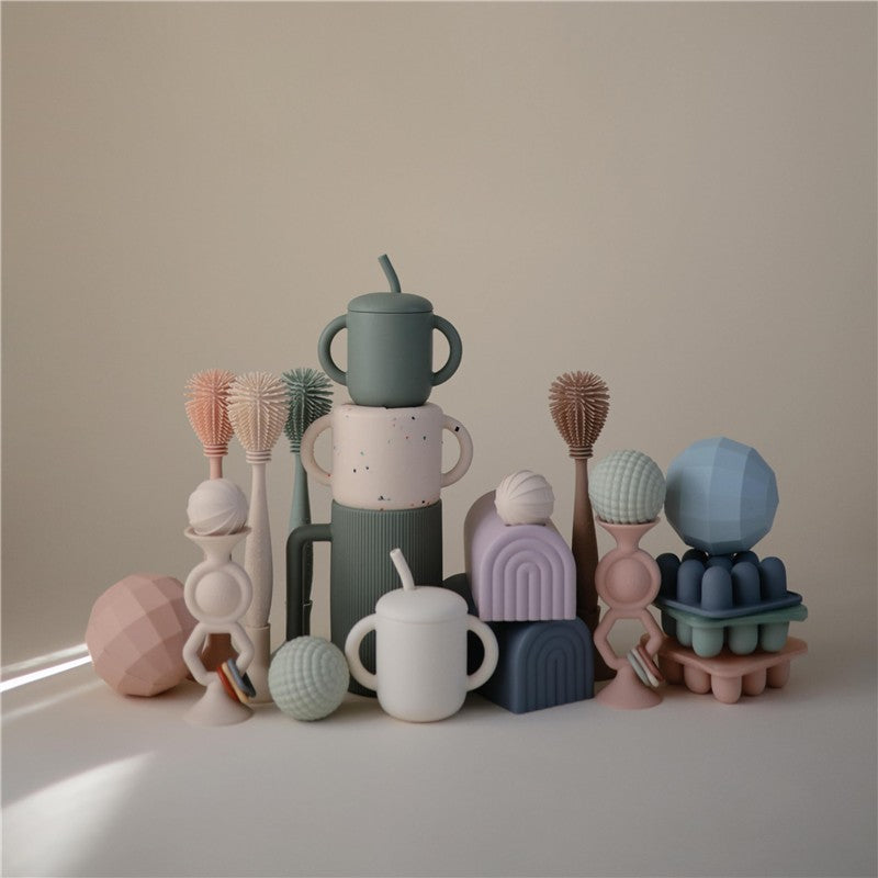 Mushie Silicone Baby Bottle Brush in blush, surrounded by colorful, playful silicone toys and shapes in various pastel colors.