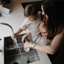 Mother and child washing a Mushie Silicone Baby Bottle Brush in a sink, water running from the faucet.