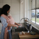 Mom wearing a pink wrap holds a baby while washing a bottle with a green brush by a sink in a bright kitchen.