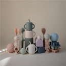 Mushie Silicone Baby Bottle Brush in Shifting Sand color, surrounded by various playful silicone shapes and textures.