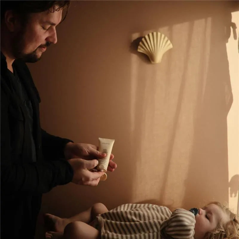 A person holds a 100 ml tube of Mushie Baby Cream, preparing to apply it to a sleeping baby dressed in striped pajamas.