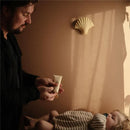 A person holds a 100 ml tube of Mushie Baby Cream, preparing to apply it to a sleeping baby dressed in striped pajamas.