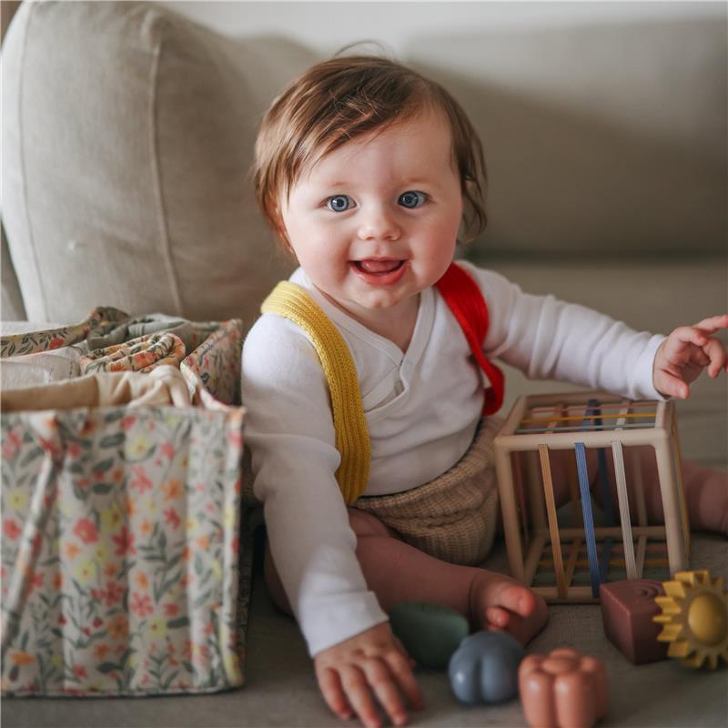 A happy baby with blue eyes sits on a couch, playing with colorful toys next to a floral-patterned diaper caddy.