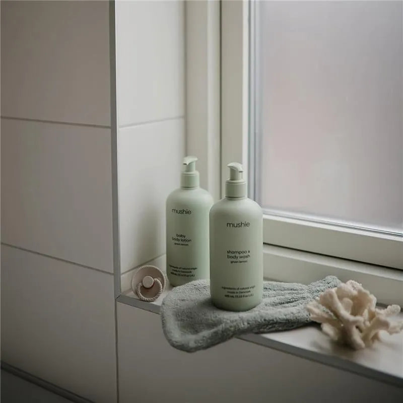 Two Mushie baby care products, a green lemon baby lotion and shampoo & body wash, are displayed on a windowsill with a soft towel.