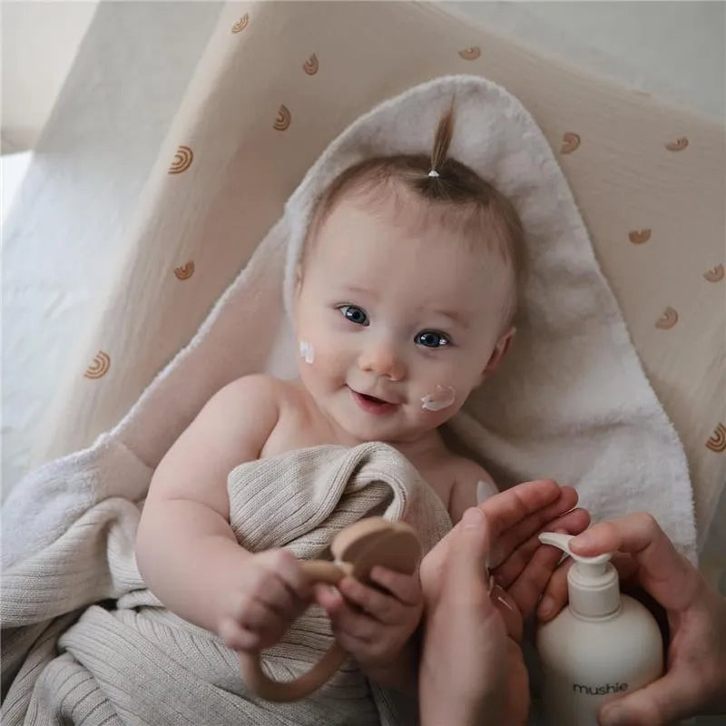A smiling baby with facial lotion sits wrapped in a cozy towel, holding a wooden toy, while a hand prepares Mushie organic body lotion.