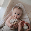 A smiling baby with facial lotion sits wrapped in a cozy towel, holding a wooden toy, while a hand prepares Mushie organic body lotion.