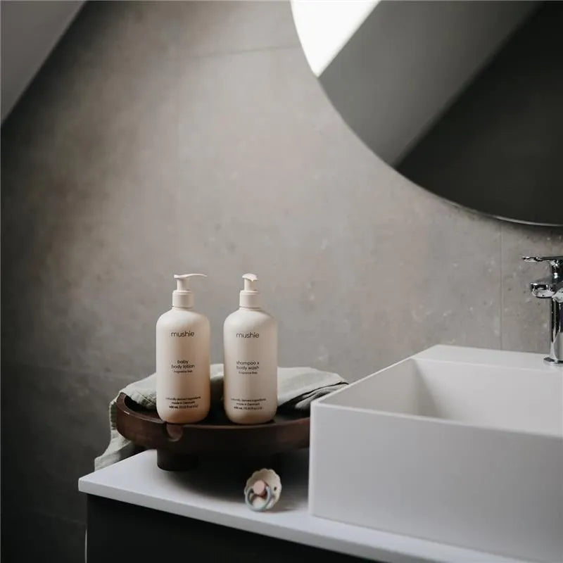Mushie organic body lotion and shampoo on a wooden tray beside a modern sink in a stylish bathroom setting.