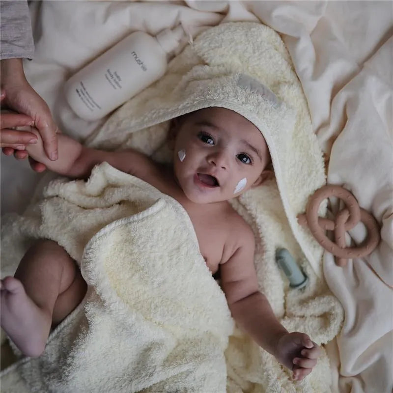 Baby wrapped in a soft yellow towel, smiling, with cream on cheeks, holding a hand, and surrounded by body lotion and toys.