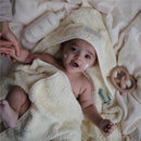 Baby wrapped in a soft yellow towel, smiling, with cream on cheeks, holding a hand, and surrounded by body lotion and toys.