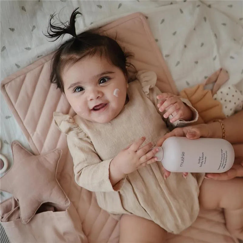 Baby smiling while holding a bottle of Mushie Baby Organic Body Lotion, with a lavender-scented cream applied on her cheek.