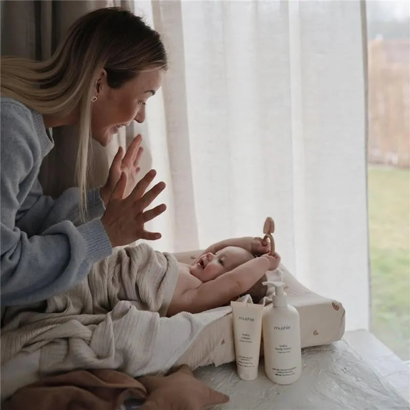 A mother joyfully interacts with her wrapped baby on a changing table, with Mushie baby lotion products nearby.