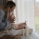 A mother joyfully interacts with her wrapped baby on a changing table, with Mushie baby lotion products nearby.