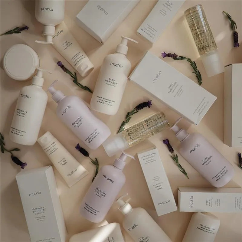 Mushie Baby Organic Body Lotion in a lavender bottle among various other baby care products on a neutral background.