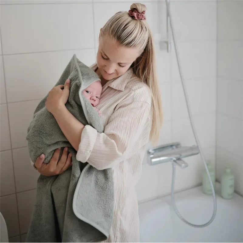 Mother holding a wrapped baby in a bathroom, with visible baby shampoo and body wash bottles in the background.