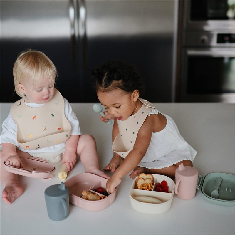 Two toddlers are seated at a table with various foods in colorful Mushie bento lunch boxes, one is blush pink.