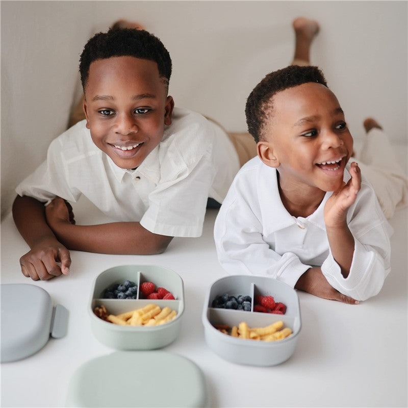 Two smiling boys lay on a white surface, showcasing the Mushie Bento Lunch Box in cloud color filled with fruit and snacks.