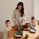 Mushie Bento Lunch Box in cloud color with fruits and a wooden bowl on a table, as a mother prepares food for two children.