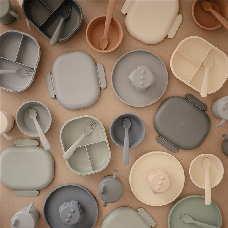 Mushie Bento Lunch Box in sage with various matching silicone dishes, utensils, and a playful figure on display.