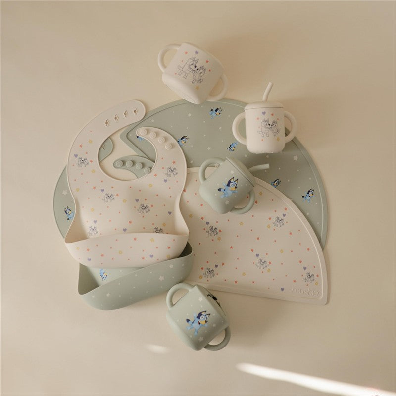 Silicone baby bib and cups in light colors with playful Bluey character designs, arranged on a soft background.