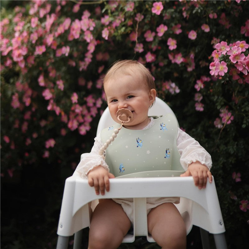 Silicone baby bib in blue with cartoon designs, worn by a seated baby in a high chair, surrounded by pink flowers.