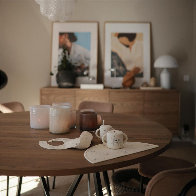 Silicone baby bib in soft sage color rests on a wooden dining table near decorative candles and ceramic mugs.
