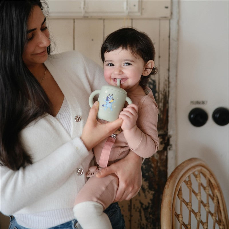 Child smiling while holding a bluey sage Mushie silicone training cup with a straw, loved by the caregiver.