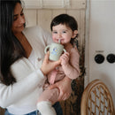 Child smiling while holding a bluey sage Mushie silicone training cup with a straw, loved by the caregiver.