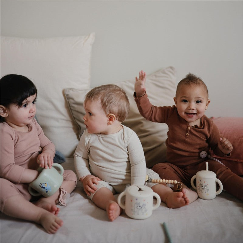 Three toddlers sitting on a bed, each holding a silicone training cup, smiling and interacting with one another.