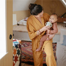 A woman in a mustard robe holds a baby in a brown outfit, who is drinking from a light blue silicone training cup with a straw.