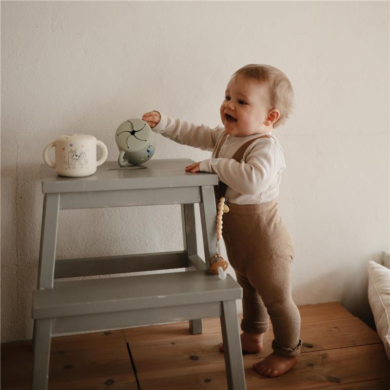 A toddler joyfully interacting with a green Bluey X Mushie Snack Cup on a wooden stool, beside a lighter cup with a cartoon design.