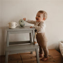 A toddler joyfully interacting with a green Bluey X Mushie Snack Cup on a wooden stool, beside a lighter cup with a cartoon design.
